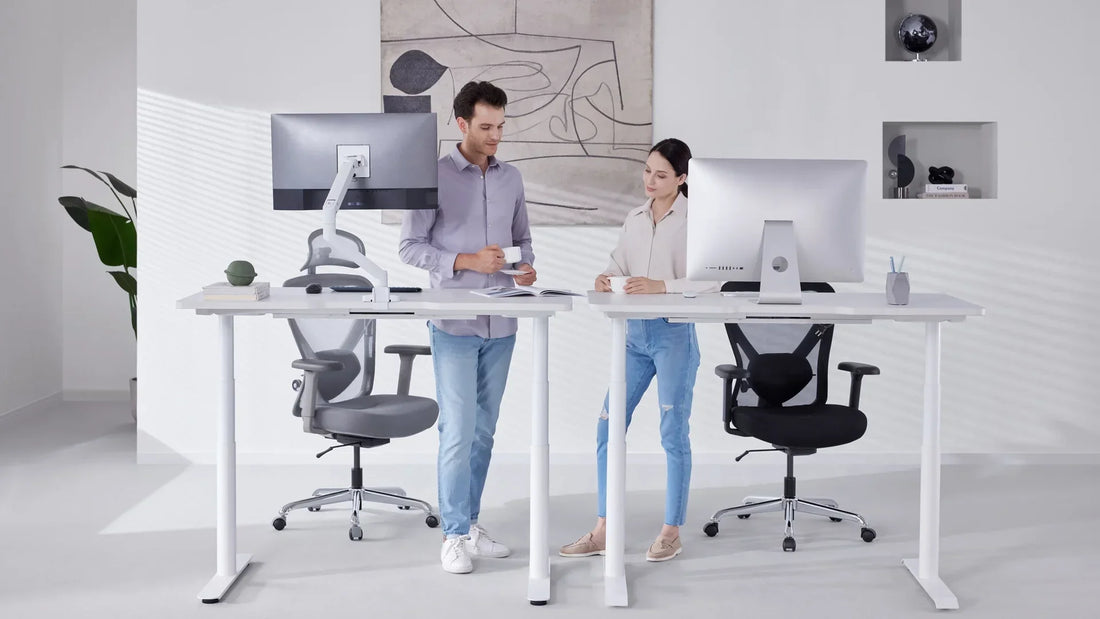 Two professionals having a discussion over coffee at a modern height-adjustable standing desk, with ergonomic office chairs positioned nearby in a well-lit workspace.