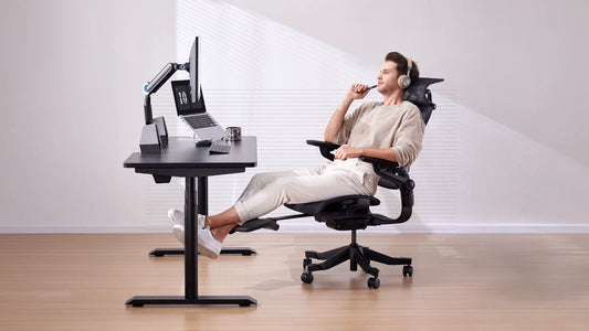 Man reclining in a Hinomi H1 Pro V2 ergonomic chair with a footrest extended, wearing headphones and working at a modern desk setup.