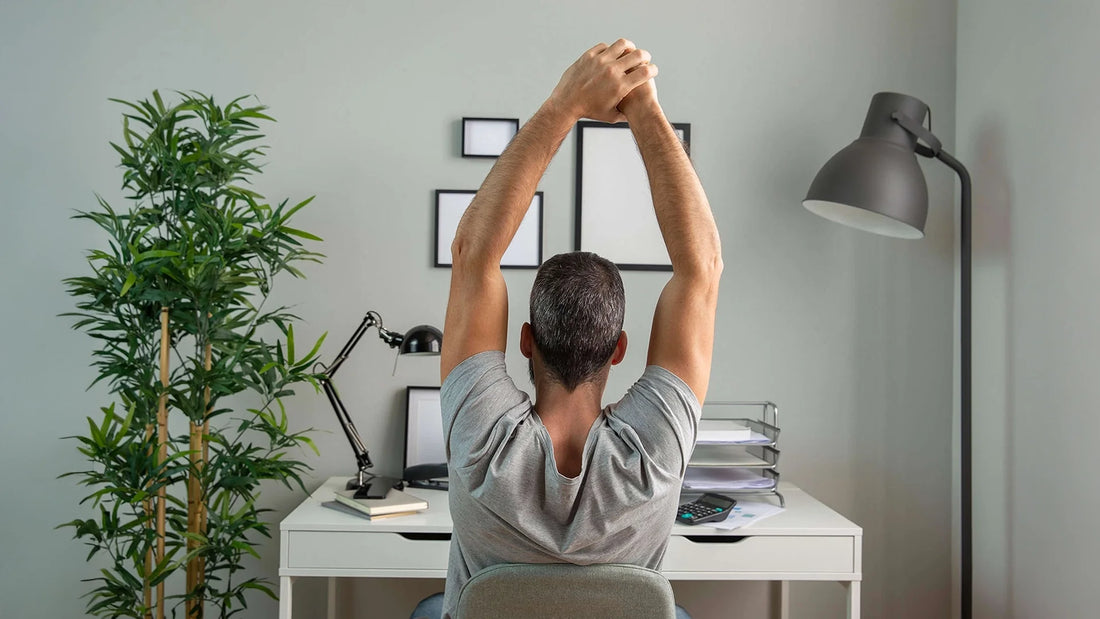 Man stretching his arms above his head while sitting at a desk in a home office, promoting ergonomic posture and workplace wellness.