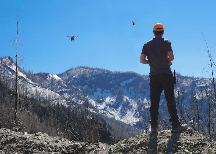 Man in an orange cap operating drones in a mountainous landscape with snow-covered peaks and a clear blue sky.